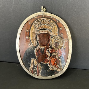 May include: An oval-shaped wooden plaque featuring a painted icon of the Black Madonna and Child. The figures wear ornate crowns and dark robes with gold-toned accents. The plaque is encased in a silver-colored metal frame with a small hanging loop.