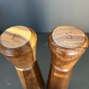 Pu&ograve; includere: Un set di due macinini per sale e pepe in legno con finitura marrone scuro. I macinini sono incisi con le lettere "P" e "S" sulla parte superiore.