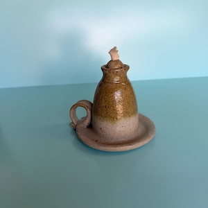 May include: A small, handmade ceramic oil lamp with a speckled, two-tone glaze. The base is a light, sandy beige, while the upper portion features a warm, golden-brown finish. It includes a small side handle and a central wick protruding from the top.
