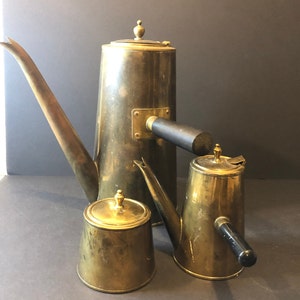 Peut inclure: Un ensemble de cafetières anciennes en laiton et un petit récipient avec des couvercles. Les cafetières ont de longs becs et des poignées noires. L'ensemble est présenté sur une surface sombre.
