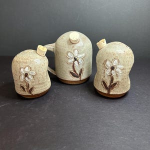 Könnte beinhalten: Ein Set aus drei Salz- und Pfefferstreuern aus Keramik mit Korkverschlüssen. Die Streuer sind mit einem weißen Blumendesign auf beigefarbenem Hintergrund verziert.