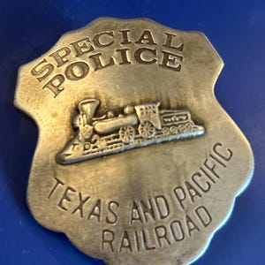 May include: A silver-colored Special Police badge from the Texas and Pacific Railroad. The badge features an embossed steam train and the words "SPECIAL POLICE" and "TEXAS AND PACIFIC RAILROAD" in a shield shape.