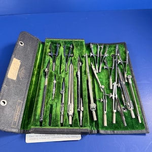 May include: A vintage drafting instrument set in a black folding case with a green velvet interior. The set includes various metal compasses, dividers, and pens. A label on the case reads "Used by R.H. Proctor, Jr. in mechanical drawing class at Hempstead H.S. 1925."