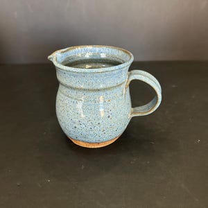 May include: A small, blue ceramic jug with a speckled glaze and a brown rim. The jug has a rounded body and a handle.
