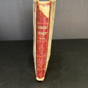 Peut inclure: Un livre vintage intitulé "Elementary Arithmetic" de White. Le livre a une reliure en cuir rouge avec des lettres dorées et des motifs décoratifs. La couverture est usée et décolorée.