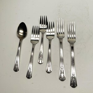 Peut inclure: Ensemble de couverts anciens en argent, comprenant des fourchettes et une cuillère, disposés sur une surface blanche. Les couverts présentent une finition argentée avec des motifs floraux décoratifs sur les manches. Les fourchettes ont quatre dents.