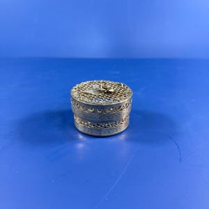 Puede incluir: Una pequeña caja redonda de metal plateado con un intrincado patrón de filigrana en los lados y la tapa. La tapa está rematada con un motivo decorativo de rosa y hoja de metal, mostrando un diseño calado detallado en toda la pieza.