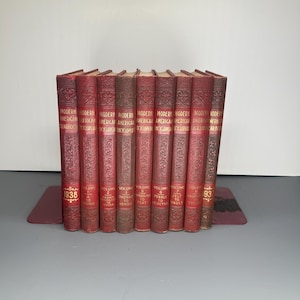 Peut inclure: Une collection de livres anciens à couverture rigide rouge, intitulée "Modern American Encyclopedia", avec des lettres dorées. Les livres sont disposés verticalement, les dos tournés vers l'avant. L'année "1938" est visible sur un volume. Les livres sont sur un serre-livres rouge.