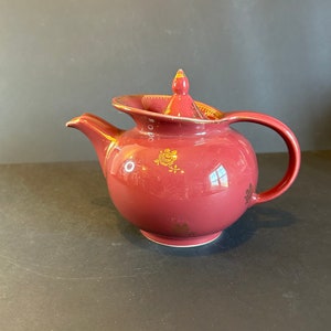 May include: A maroon teapot with a gold lid and floral design. The teapot has a round body and a curved spout.