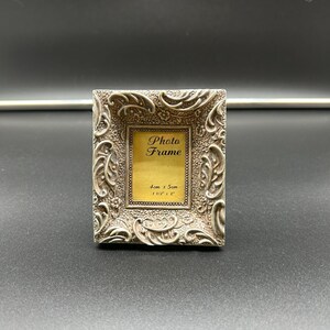 May include: A small, ornate silver photo frame with a rectangular opening. The frame is decorated with swirling patterns and the text "Photo Frame" is visible. The frame dimensions are 1 1/2" x 2".