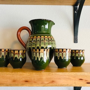 May include: A green ceramic pitcher with a brown handle and six matching cups. The pitcher and cups have a decorative pattern of wavy lines in shades of brown, yellow, and green. The set is displayed on a wooden shelf.