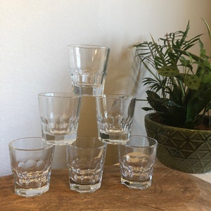 Puede incluir: Una pirámide de siete vasos de chupito transparentes y facetados. Los vasos están apilados sobre una superficie de madera, con una planta verde en maceta en el fondo. Los vasos tienen un diseño clásico y robusto.