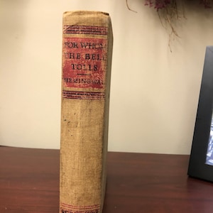 以下が含まれることがあります： 茶色の布張りのカバーと赤と黒のストライプが特徴のヴィンテージハードカバーの本。タイトル「誰がために鐘は鳴る」と著者「ヘミングウェイ」は、赤い背景に黒で印刷されています。出版社「スクリブナー」は、背表紙の下部に赤い背景に黒で印刷されています。