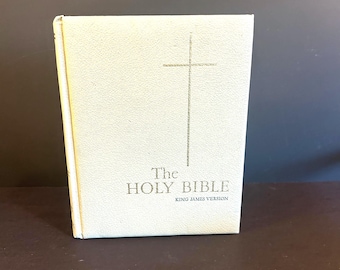 May include: A cream-colored book with the title "The Holy Bible" and a cross design. The text "King James Version" is below the title. The book has a textured cover and a gold spine.