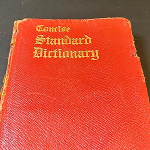 Peut inclure: Un livre rouge vintage, "Concise Standard Dictionary", avec des lettres dorées. La couverture du livre est texturée et montre des signes d'usure. Le titre est affiché en évidence dans une police ornée.