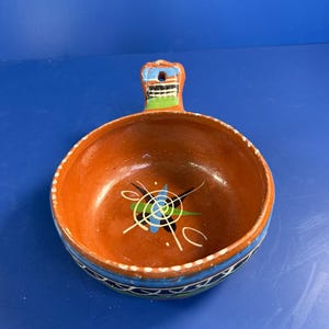 May include: A round terracotta bowl with a single handle featuring a hole. The interior is reddish-brown with a central painted star-like design in blue, green, and white. The exterior rim has a blue band with white geometric patterns.