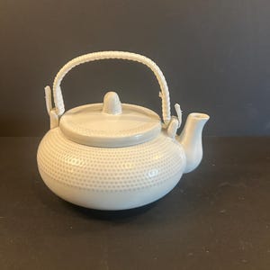 Può includere: Una teiera in ceramica bianca con un motivo strutturato e puntinato. La teiera ha un beccuccio curvo, un coperchio con un piccolo pomello e un manico intrecciato. La teiera è progettata per preparare il tè.