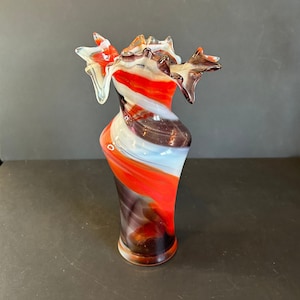 Może przedstawiać: Szklany wazon z unikalnym, abstrakcyjnym wzorem. Wazon wykonany jest z przezroczystego szkła z czerwonymi, białymi i czarnymi wirami. Górna część wazonu jest rozszerzona i ma falowany brzeg.