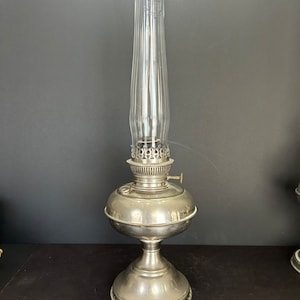 Könnte beinhalten: Eine silberne Petroleumlampe mit einem klaren Glaszylinder. Die Lampe hat einen runden Sockel mit einem dekorativen Rand und einen runden Korpus mit einem dekorativen Oberteil.