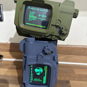 May include: A 3D printed replica of a Pip-Boy from the Fallout video game series. The Pip-Boy is a device worn on the wrist that displays information about the player's character, inventory, and the game world. The Pip-Boy is green and has a black screen with green text. The Pip-Boy is sitting on top of a grey 3D printed replica of a Pip-Boy.