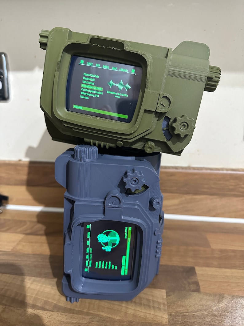 May include: A 3D printed replica of a Pip-Boy from the Fallout video game series. The Pip-Boy is a device worn on the wrist that displays information about the player's character, inventory, and the game world. The Pip-Boy is green and has a black screen with green text. The Pip-Boy is sitting on top of a grey 3D printed replica of a Pip-Boy.