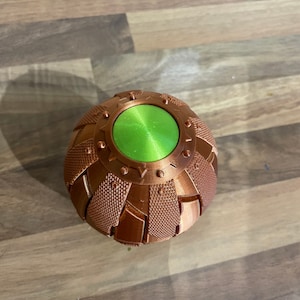 May include: A copper-coloured, spherical object with a green, circular button on top. The object has a textured surface and appears to be made of metal.