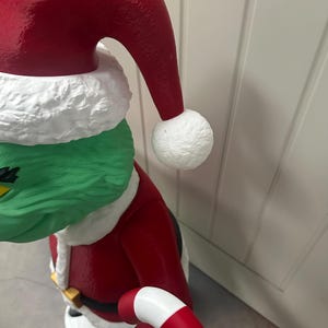May include: A green character wearing a red Santa Claus suit and hat with white trim and a white pom-pom. The character is holding a red and white candy cane. The figure has a green face and is wearing a black belt with a gold buckle.