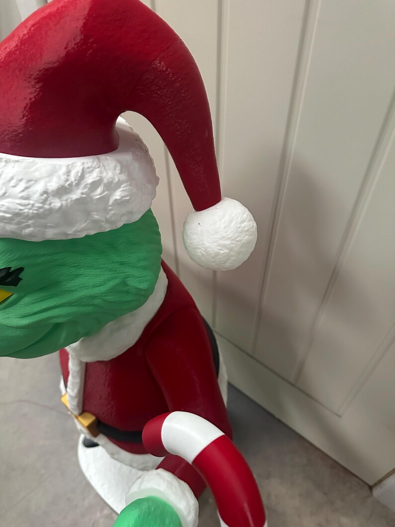 May include: A green character wearing a red Santa Claus suit and hat with white trim and a white pom-pom. The character is holding a red and white candy cane. The figure has a green face and is wearing a black belt with a gold buckle.