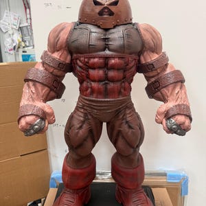 May include: A detailed, full-body statue of the Juggernaut, a Marvel Comics character. The figure is predominantly red and brown, with a helmet, armor, and muscular physique. The statue is standing on a black surface.