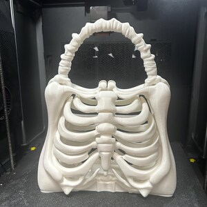 May include: A white, 3D-printed handbag in the shape of a human rib cage. The bag has a handle designed to look like a spine. The skeletal design is detailed. The bag is on a dark surface.