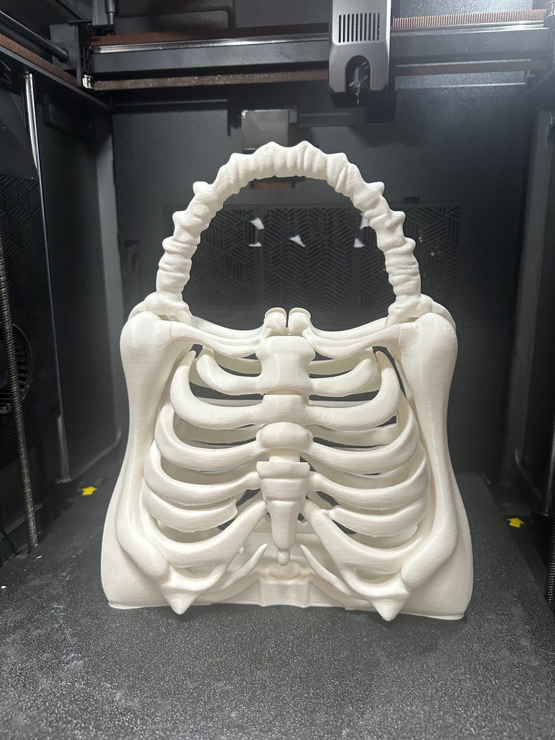 May include: A white, 3D-printed handbag in the shape of a human rib cage. The bag has a handle designed to look like a spine. The skeletal design is detailed. The bag is on a dark surface.