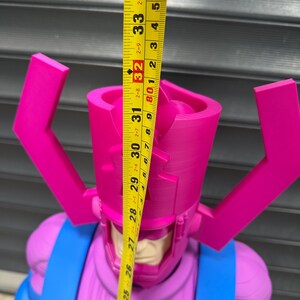 May include: A large, pink and blue action figure of a character with a helmet and antenna-like structures. The figure is approximately 32 inches tall, as measured by a yellow tape measure. The figure has a blue torso and pink head.