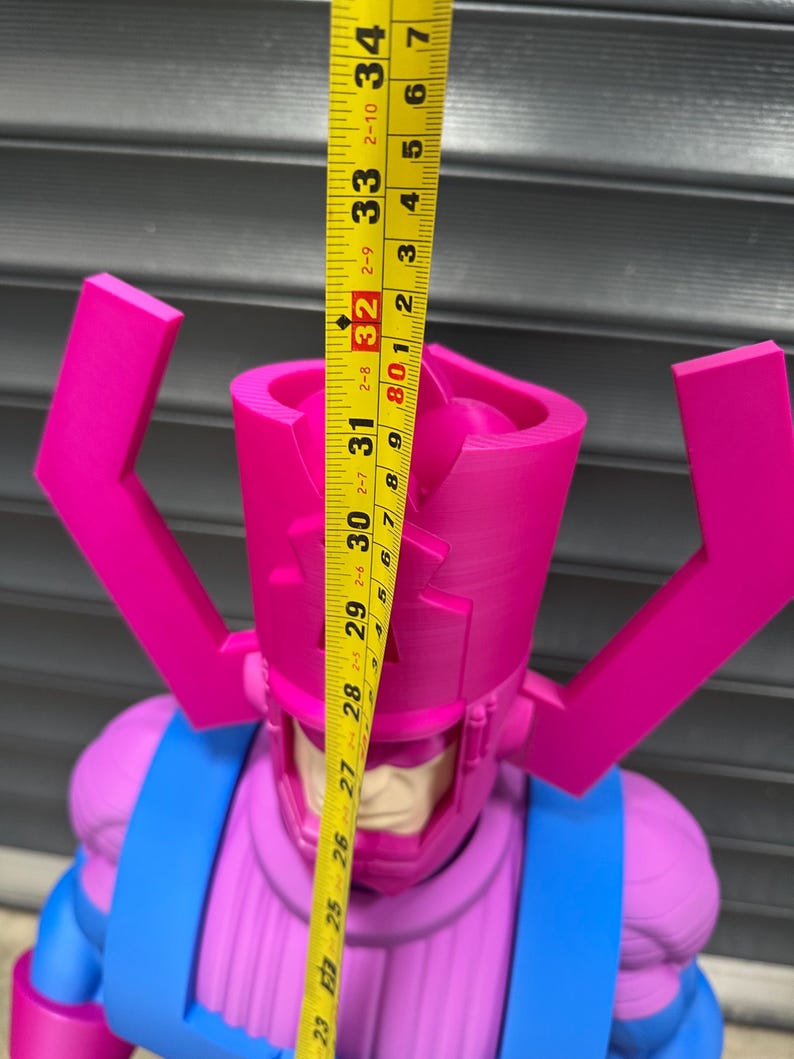 May include: A large, pink and blue action figure of a character with a helmet and antenna-like structures. The figure is approximately 32 inches tall, as measured by a yellow tape measure. The figure has a blue torso and pink head.