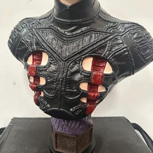 May include: A detailed bust of a character with a pale face, nails in the head, and a stitched mouth. The figure wears a black leather-like garment with red accents. The base is a dark color with a purple and brown design.