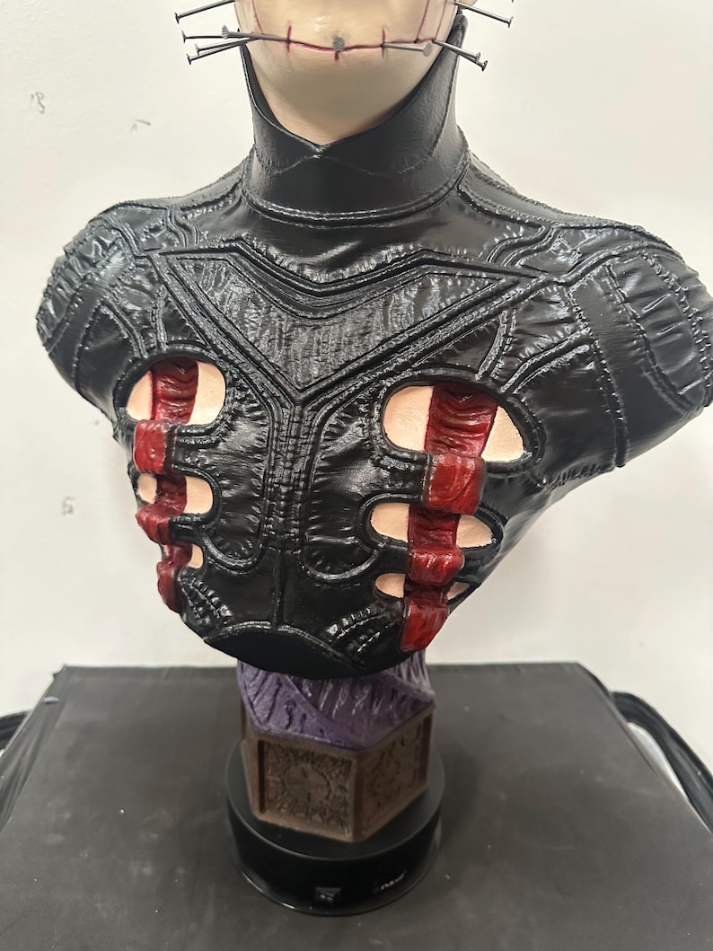 May include: A detailed bust of a character with a pale face, nails in the head, and a stitched mouth. The figure wears a black leather-like garment with red accents. The base is a dark color with a purple and brown design.