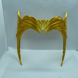 May include: A gold-coloured, ornate headband with a decorative, wave-like design. The headband is made of a smooth, shiny material and has a slightly curved shape.