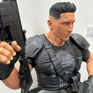 May include: A detailed bust of a character in a black tactical outfit, holding two black rifles. The figure has short, dark hair and a stern expression. The character's shirt features a white skull emblem.
