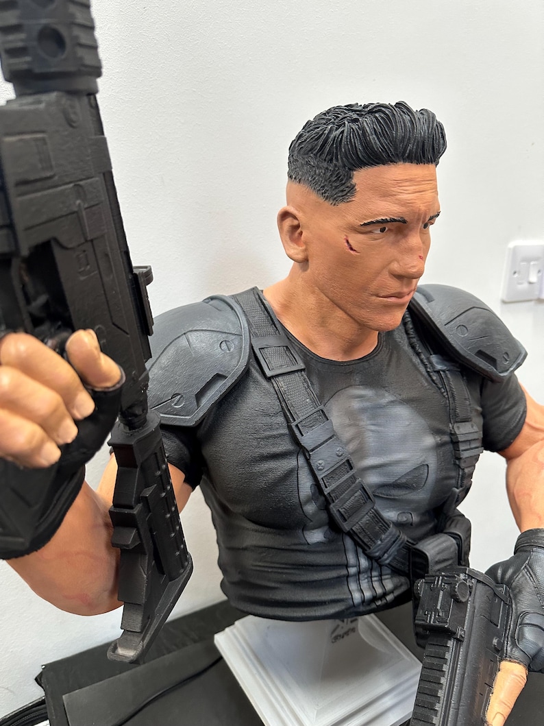 May include: A detailed bust of a character in a black tactical outfit, holding two black rifles. The figure has short, dark hair and a stern expression. The character's shirt features a white skull emblem.