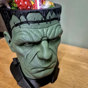 Puede incluir: Un recipiente verde con forma de cabeza de monstruo de Frankenstein lleno de caramelos envueltos de colores. El recipiente tiene detalles de costura negros y un cuello negro. Una divertida y espeluznante decoraci&oacute;n de Halloween.