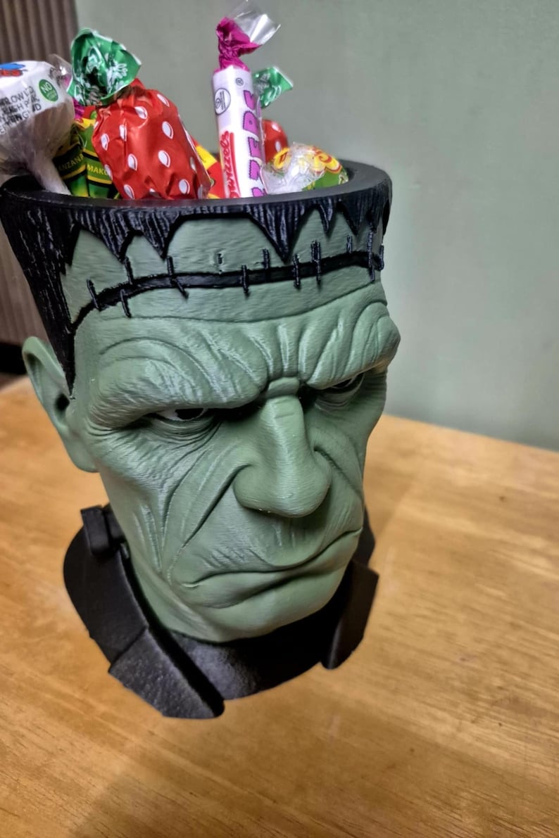 Puede incluir: Un recipiente verde con forma de cabeza de monstruo de Frankenstein lleno de caramelos envueltos de colores. El recipiente tiene detalles de costura negros y un cuello negro. Una divertida y espeluznante decoraci&oacute;n de Halloween.