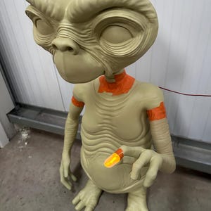 May include: A beige-colored alien figure with large eyes and a distinctive head shape. The figure has orange accents on the neck and arms, and is holding a glowing orange object. The figure is a representation of the character E.T.