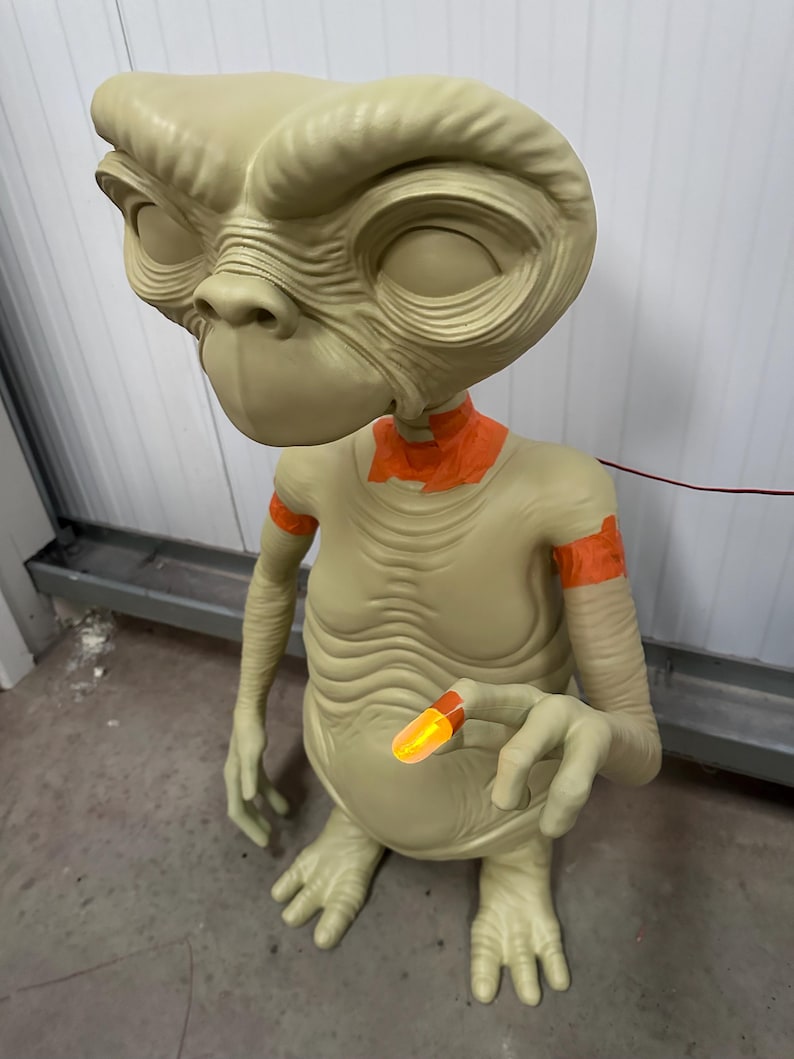 May include: A beige-colored alien figure with large eyes and a distinctive head shape. The figure has orange accents on the neck and arms, and is holding a glowing orange object. The figure is a representation of the character E.T.