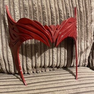 May include: A red plastic headband with a detailed, intricate design. The headband is shaped like a crown with two points extending upwards.