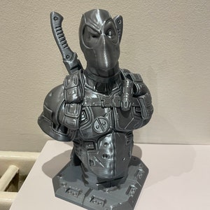 May include: A gray 3D printed bust of Deadpool, a superhero character, with a sword on his back. The bust is on a gray base.