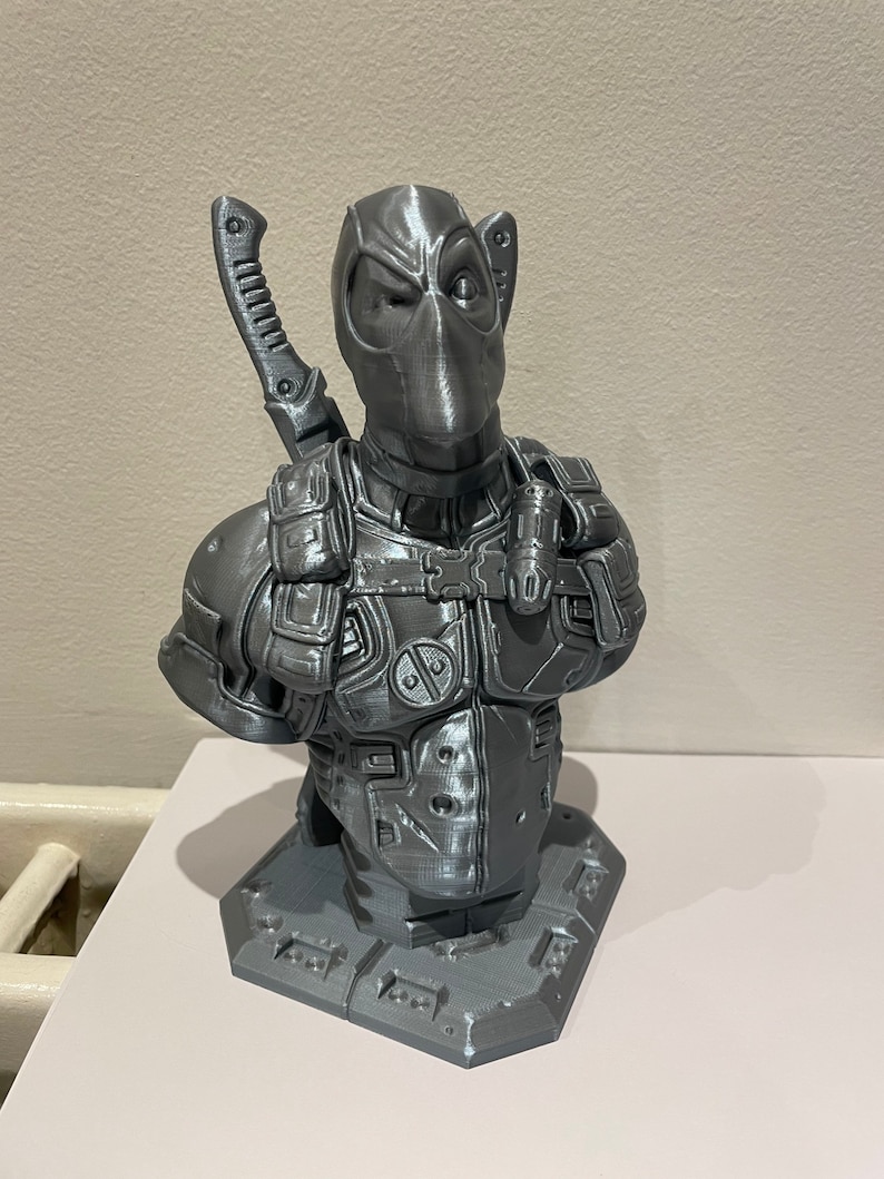 May include: A gray 3D printed bust of Deadpool, a superhero character, with a sword on his back. The bust is on a gray base.