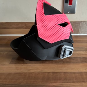 May include: A black and pink 3D printed helmet with a textured design. The helmet has a visor and a small, gray, rectangular detail on the side.