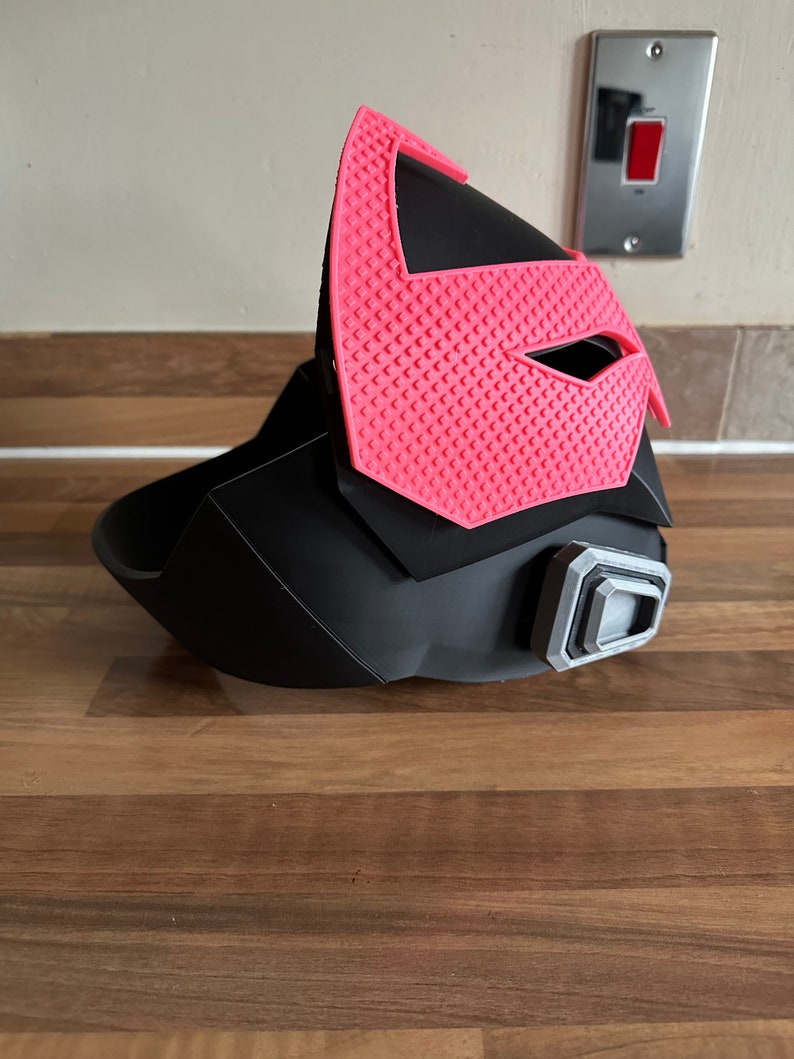 May include: A black and pink 3D printed helmet with a textured design. The helmet has a visor and a small, gray, rectangular detail on the side.