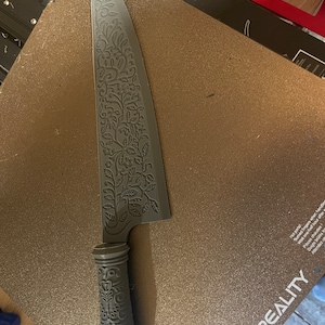 Può includere: Un coltello grigio stampato in 3D con un design floreale dettagliato sulla lama e sul manico. Il coltello è appoggiato su una superficie marrone testurizzata con la scritta "CREALITY" stampata su di essa.