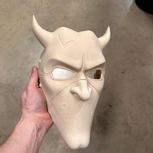 May include: A beige mask with a devilish design, featuring curved horns, a pointed nose, and a stern expression. The mask has eye openings and is likely made of a rigid material. The mask is held by a person.