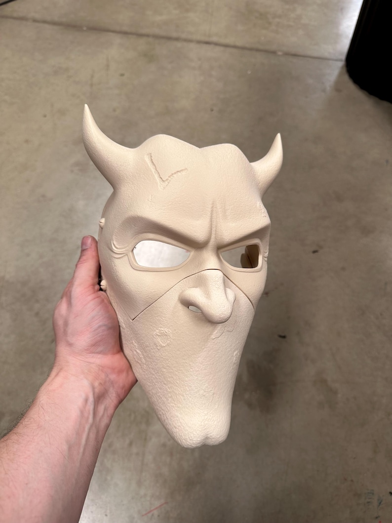 May include: A beige mask with a devilish design, featuring curved horns, a pointed nose, and a stern expression. The mask has eye openings and is likely made of a rigid material. The mask is held by a person.